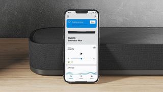 three quarter image showing Sennheiser Ambeo Soundbar Plus on wooden surface with app displayed on smartphone