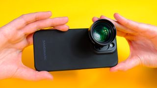 A photo of a Sandmarc iPhone lens attached to an iPhone 15 Pro using the Sandmarc iPhone case, all against a yellow background