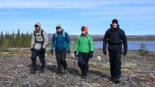 four people in rugged outdoor gear walk beside a remote lake in the canadian wilderness