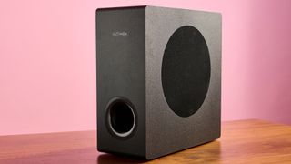 Ultimea Nova S50 subwoofer on a wooden surface against a pink background