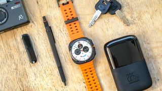 Samsung Galaxy Watch Ultra on an orange strap sitting on a wood table next to small accessories like keys, a pen and a pocket knife