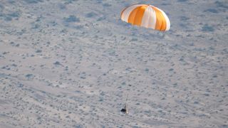 a small capsule descends toward a desert landscape beneath an orange and white parachute.