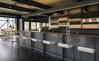 Left: Seating area with high tables and chairs with a vase of flowers in the center. Right: The restaurant's grey coloured bar with a display of wine bottles and 6 white bar stools