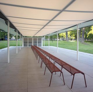 White frame and cream roof Pavilion with white flagged floor, line of red metal benches, surrounded by grass and trees in the distance