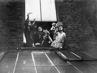 a group of 6 people lean out of a window and hold dark film up to the sky so they can view the solar eclipse through it.