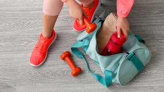 Someone packing a gym bag on the floor with dumbbells, a water bottle and a towel