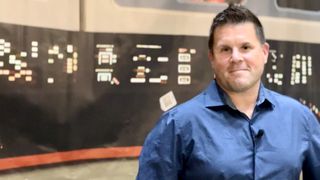 a man in a blue collared shirt stands in front of a black panel with various buttons.