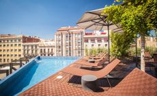 A hotel pool area on a building roof with a triangular shaped pool, wooden decking, wicker loungers, round side tables and potted trees all surrounded by buildings in a city.