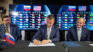 Three people in suits sit at a table. Behind them, various nations flags are featured on television screens.