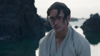 Close up of a man's face. He is standing on a beach, his chin-length hair sweeping in the breeze. He has a serious expression on his face.