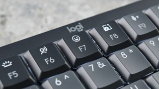 A photo of the Logitech MX Mechanical Mini keyboard in black and gray, on a stone slab and wooden table with a blue background.