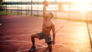 Man holding kettlebell above head with right arm and kneeling on left knee with right leg bent during outdoor kettlebell workout