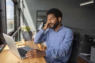 A man in a blue shirt sits at his dek with his hand placed to his head in deep concentration