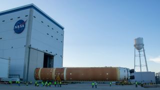 A large orange rocket booster stage lays horizontal on wheels outside a tall hangar and a water tower.