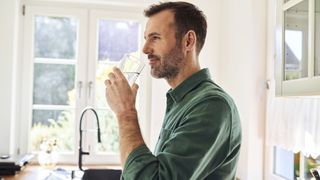 A man with short dark hair and wearing a forest green shirt drinks a glass of water in a bright and sunny kitchen on a summer's morning