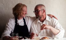 Female and male chef sat next to each other smiling at each other with food and drink on their table. 