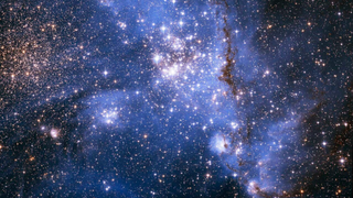 A blue scene of space with lots of stars, many of which sparkly quite brightly in the center. Streams of gas and other material can be seen as well.