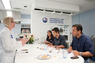 a woman in a white lab coat stands in front of three people in dark shirts seated at a white table with food and drinks.