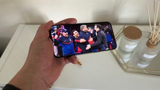 An Apple iPhone 15 Pro Max held in landscape orientation in front of a table bearing an incense burner. Ted Lasso from Apple TV+ is on the screen