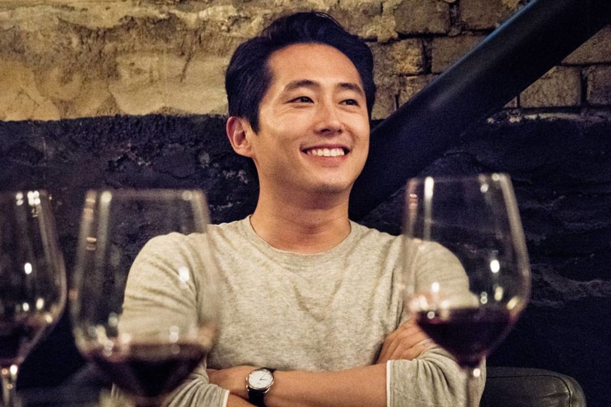 Steven Yeun wearing a white sweater and sitting at a table with wine glasses around him and a big smile on his face in Burning