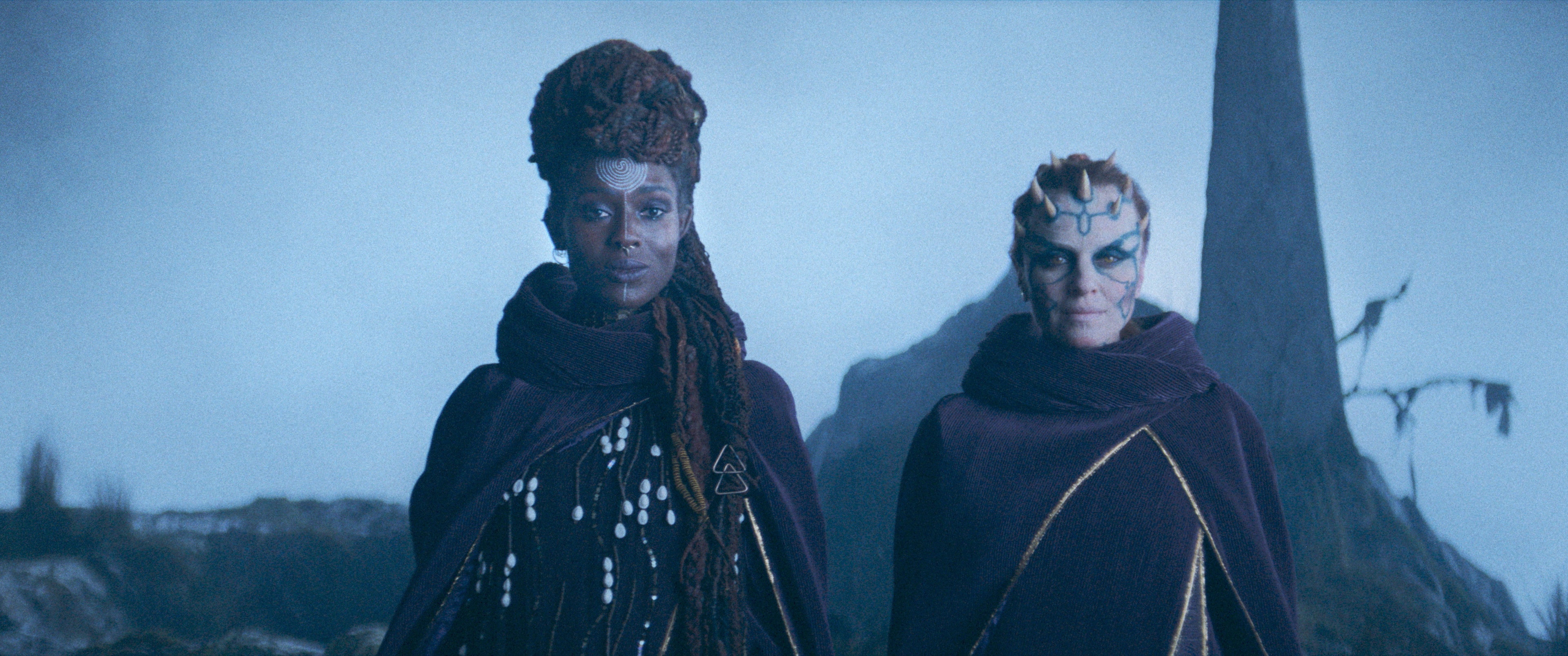 (L-R): Mother Aniseya (Jodie Turner-Smith) and Koril (Margarita Levieva) in The Acolyte. They stand abreast, smiling slightly, in elaborate robes on a stark cliff face.