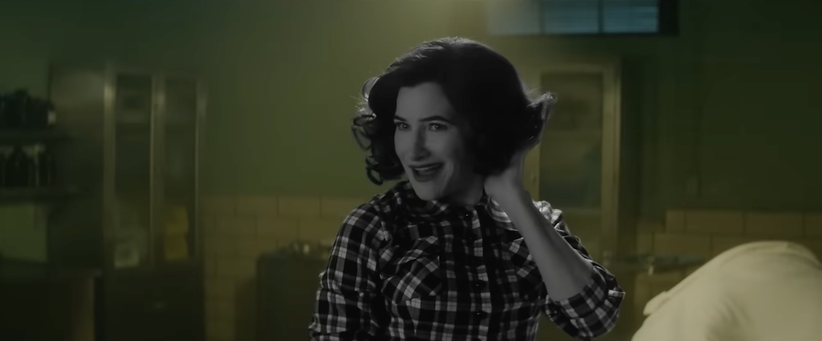 Kathryn Hahn as Agatha/Agnes in Agatha All Along. She smiles and preens her hair, standing in a green tiled room. She, however, appears entirely in black and white, like an old sitcom.