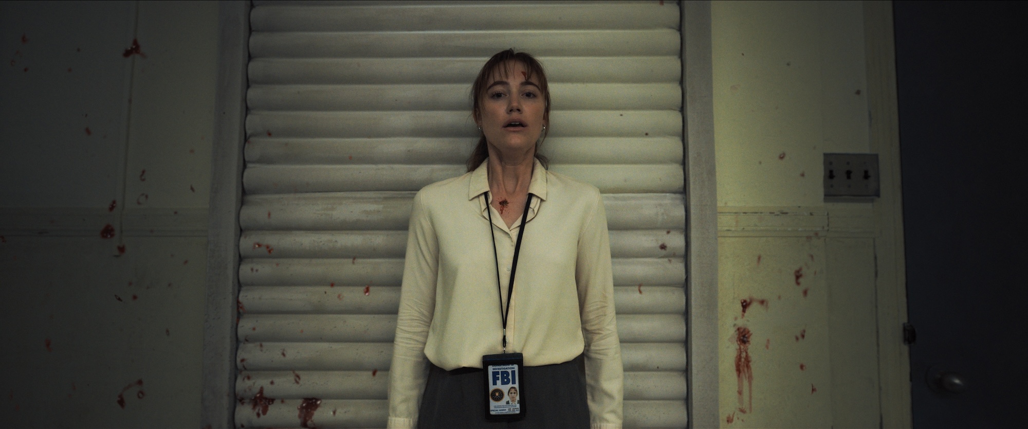 A woman wearing an FBI ID lanyard stands in what seems to be a dim, blood-splattered room in Longlegs.