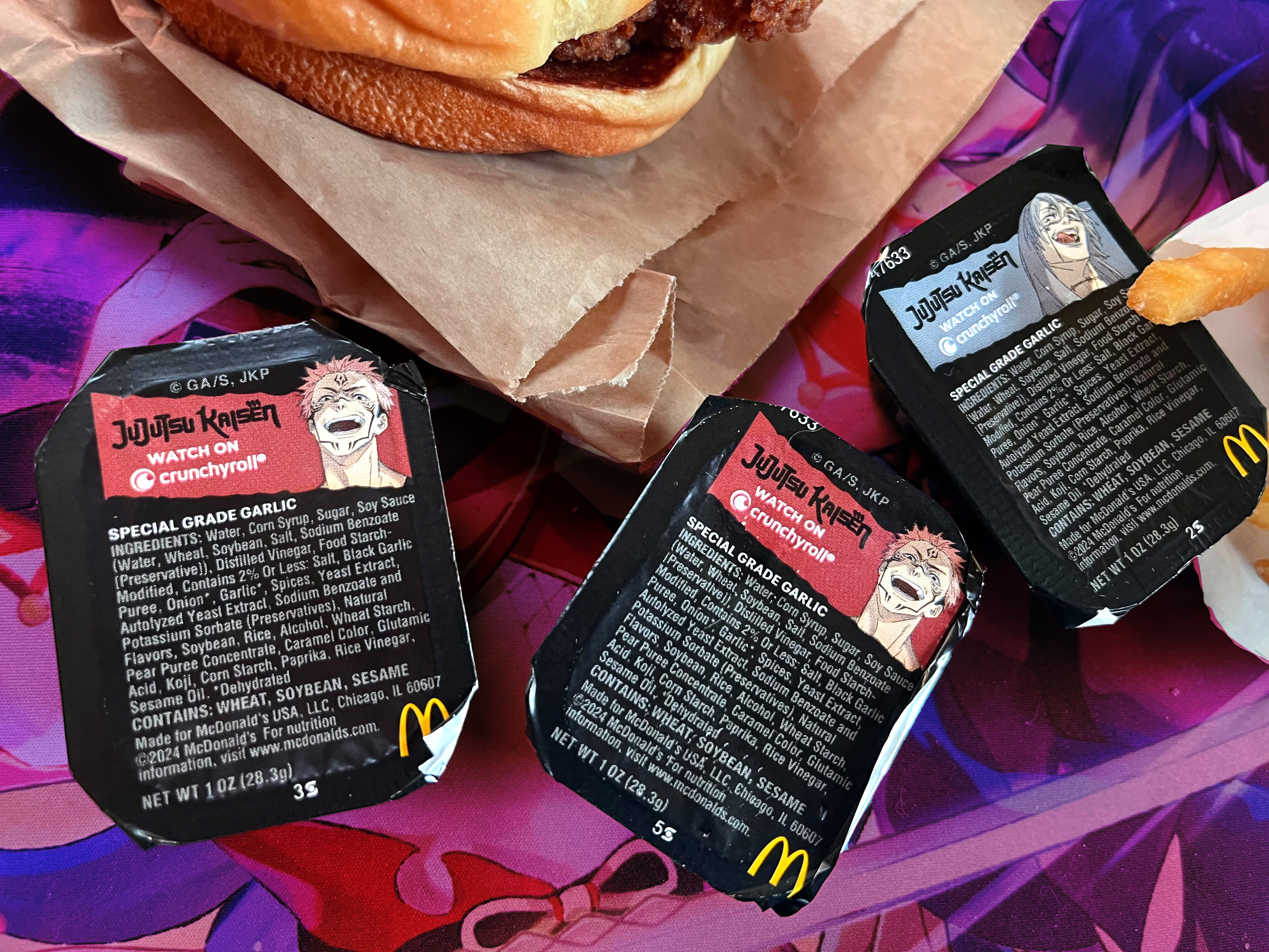 An photo of the Jujutsu Kaisen McDonald’s sauces on a desk. You can see tiny packages with writing; two have the faces of Sukuna and one has Mahito on it.