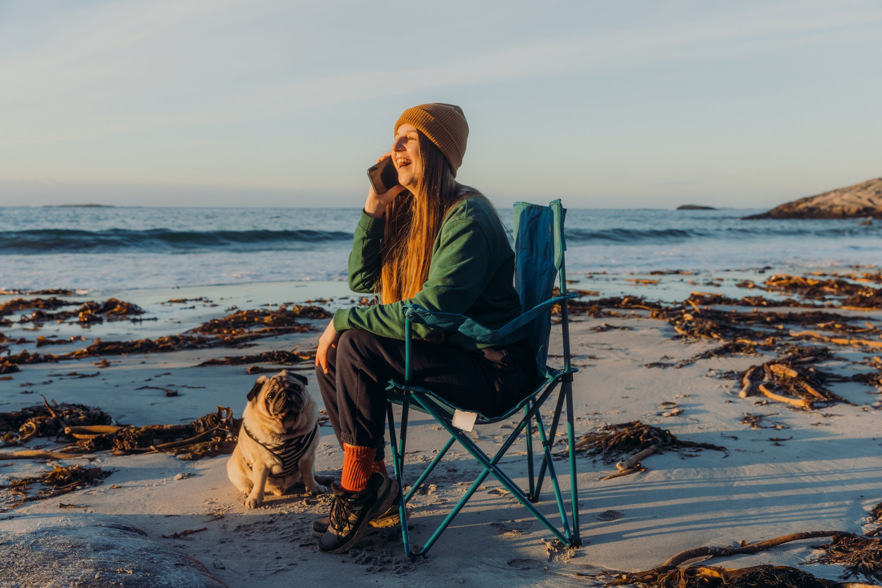 Lady sitting on the beach in a camping chair with a pug dog at her side