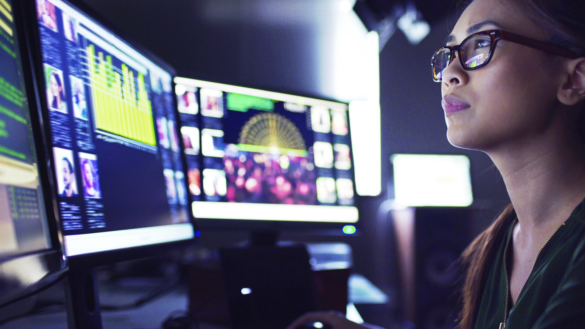 A young woman sitting down at her desk where she’s surrounded by 3 large computer monitors displaying out of focus images of people as thumbnails; crowds; graphs & scrolling text.