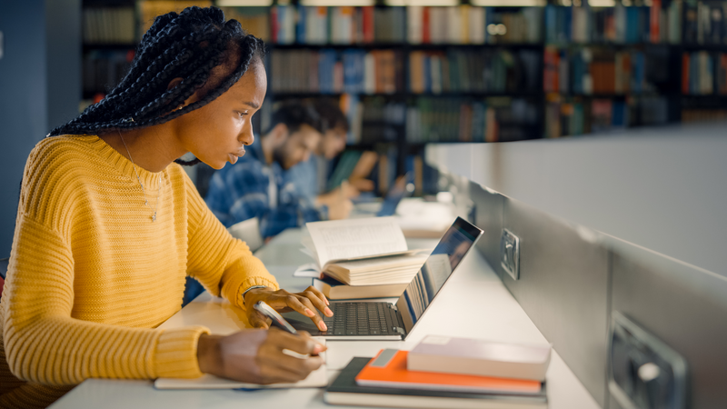 University Library: Gifted Black Girl uses Laptop, Writes Notes for the Paper, Essay, Study for Class Assignment. Diverse Multi-Ethnic Group of Students Learning, Studying for Exams, Talk in College