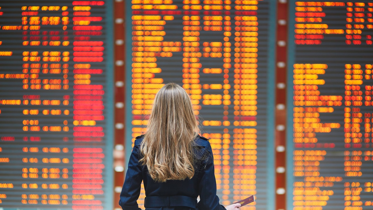 Young woman in international airport looking at the flight information board, holding passport in her hand, checking her flight