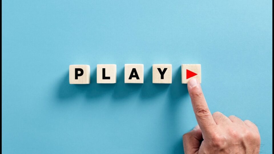 The word PLAY spelled out in wooden blocks on a blue background with a real hand and finger pressing a play button on a block at the end of the word.