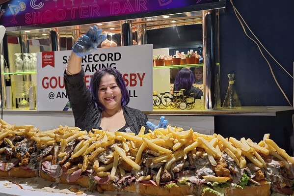 In this photo provided by Joe Redelinghuys. left, for Annies Ladies Bar, Annie Redelinghuys poses with a giant Gatsby sandwich on display at the establishment in Cape Town, South Africa, Saturday, June 29, 2024. A South African restaurant and bar has served up a 3-meter-long (9.8 feet) sandwich in the latest offering from an establishment with a taste for the supersize. (Annies Ladies Bar via AP)