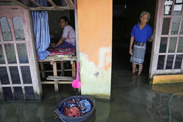 Sudarto stands at the door of his flooded home as his daughter Turiah looks on at their flooded house in Timbulsloko, Central Java, Indonesia, Sunday, July 31, 2022. With a physical disability that prevents Turiah from normal work in the village, she spends her day sitting in the home's front window on an elevated wooden platform. (AP Photo/Dita Alangkara)