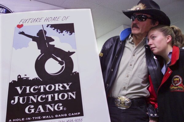FILE - Montgomery Lee Petty, 14, leans on her grandfather, racing great Richard Petty, as she listens to her dad Kyle Petty, not shown, announce that the Petty family is founding The Victory Junction Gang Camp during a news conference at the North Carolina Speedway near Rockingham, N.C., Saturday, Oct. 21, 2000. Adam Petty was 19 when he was killed in a crash practicing for a race at New Hampshire. Not too many years before, he'd made a visit to Paul Newman's-owned Camp Boggy Creek and became transfixed in creating a similar camp in North Carolina for children with serious medical issues. Richard Petty said the family following through on Adam's dream with be their lasting legacy. (AP Photo/Erik Perel, File)