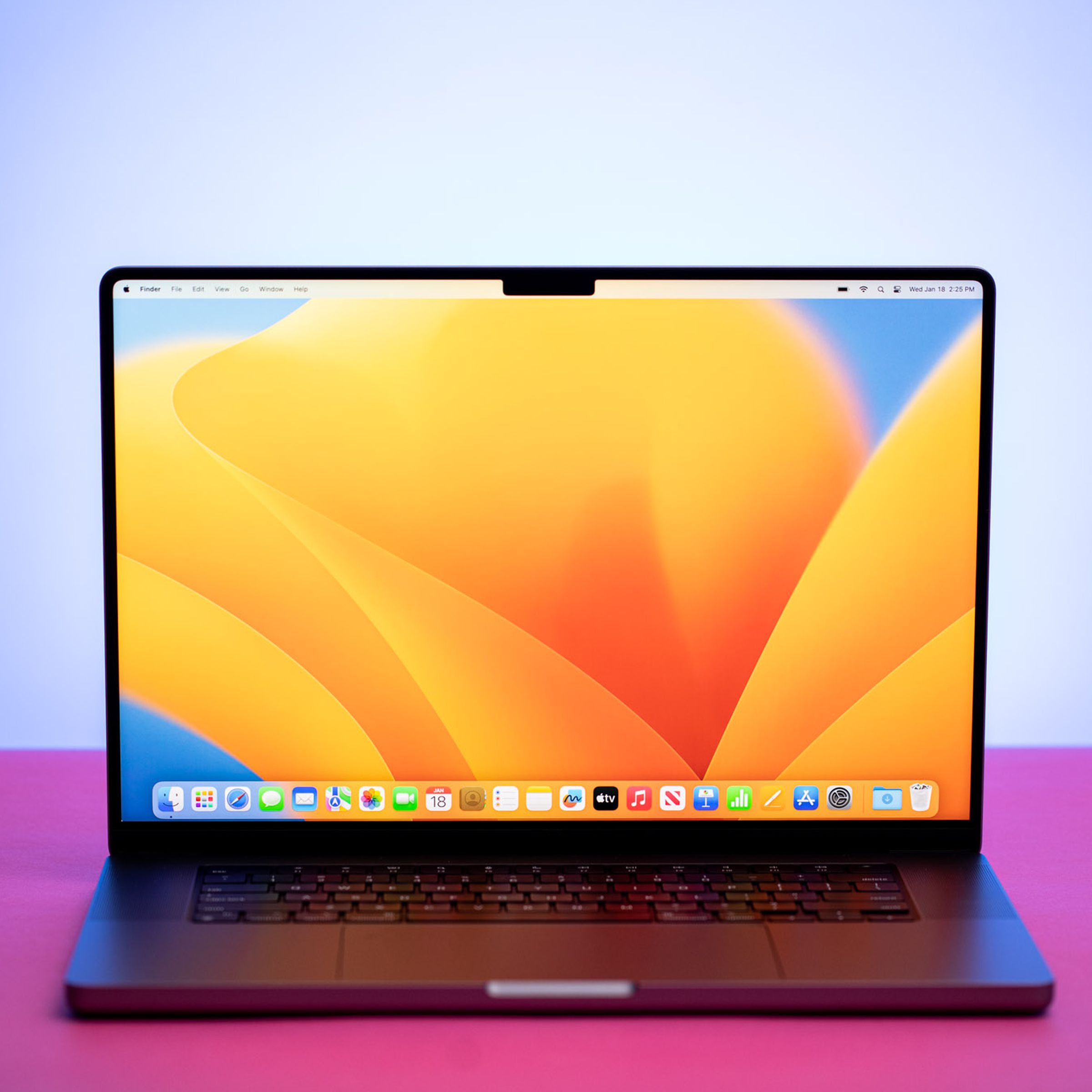 The MacBook Pro 16 (2023) on a pink table. The screen displays a blue and yellow desktop pattern.