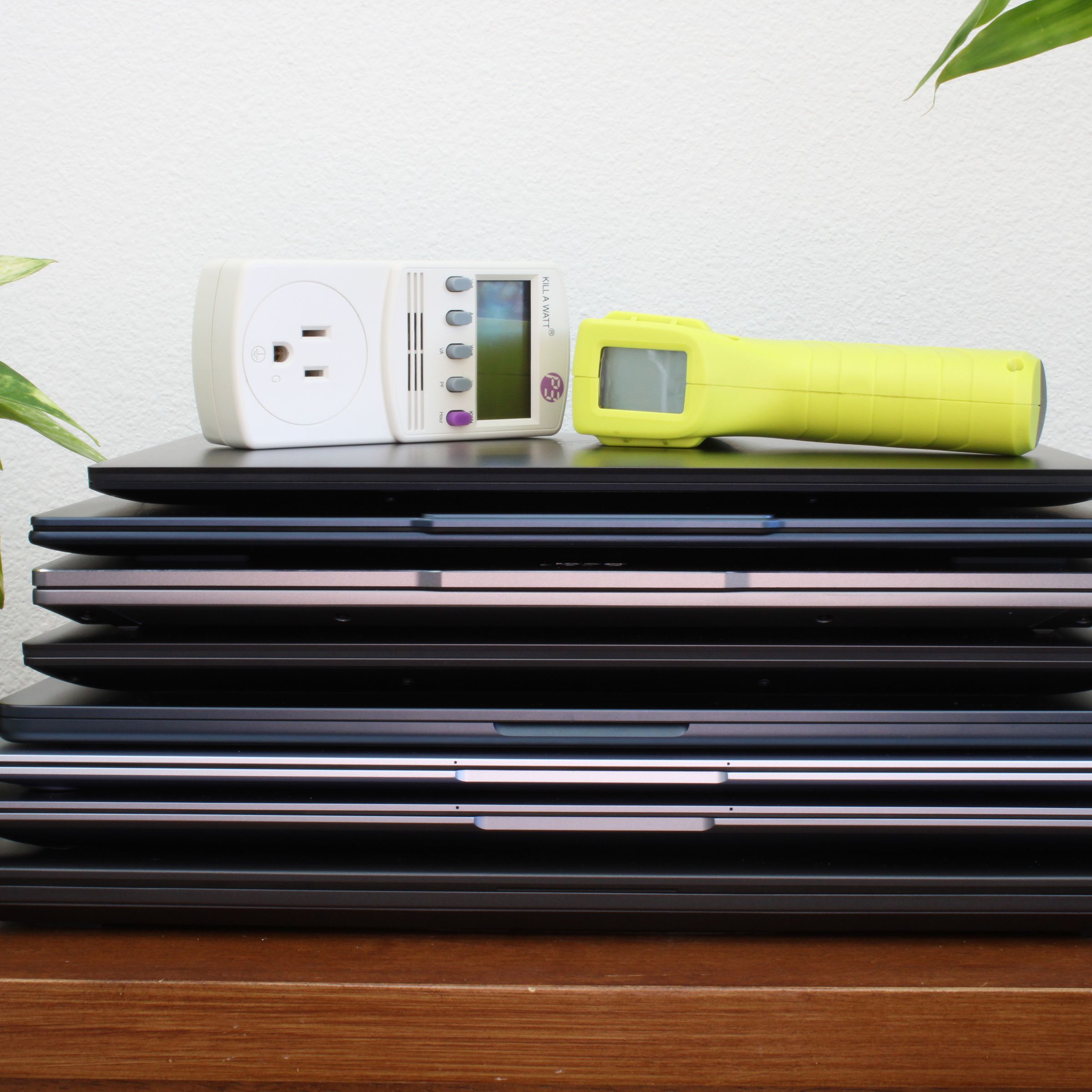 A stack of laptops on top of a brown table, flanked by two green plants, against a white wall.