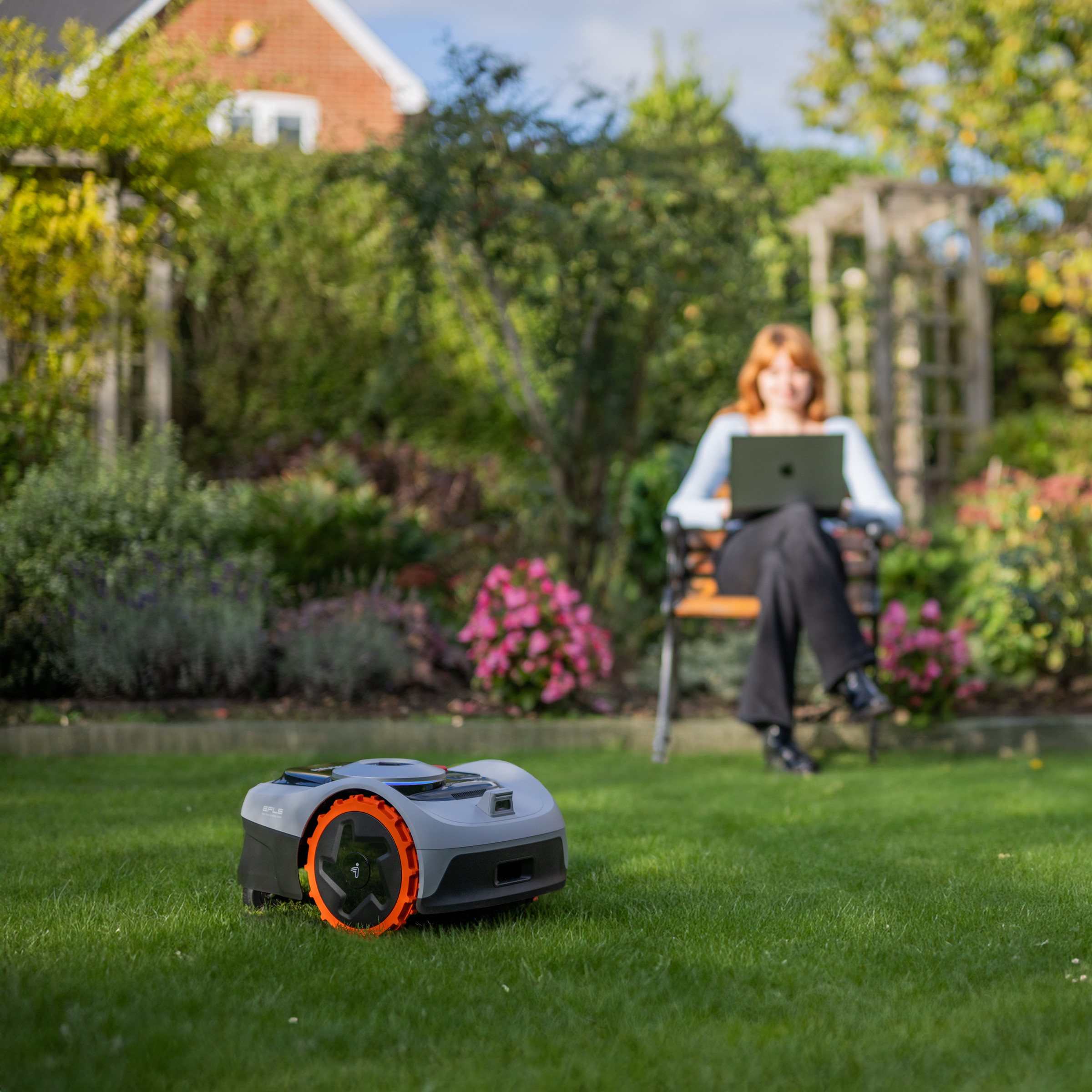 A robot lawnmower on a lawn with a lady sitting in a chair on her computer.