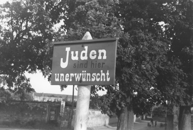 An anti-Jewish sign posted on a street in Bavaria reads "Jews are not wanted here." Julien Bryan took this photograph while visiting Germany in 1937. Back in the United States, Bryan regularly gave lectures with accompanying motion pictures to convey the looming dangers he foresaw in Europe.
During one of these presentations in 1938, he said: "And then a sign like this. Along the Rhine you see these signs against the Jew everywhere, … all through central and southern Germany, saying simply and uniformly the same thing. Jews are not wanted here…. Out of my own curiosity because I am a reporter who is anxious to get both sides of the story – I talked further with these peasants and in a number of cases I asked the German people along the Rhine … how come these signs? Who put them up? They rather laughed about it all and not too pleasantly, and they denied having anything to do with it..."
Citizens would have viewed this sign in public every day. Think about which municipal officials might have had to approve the content of the sign and its display in a public area. Who might have created it and decided where to hang it? What does this indicate about the involvement of citizens and officials in public discrimination?