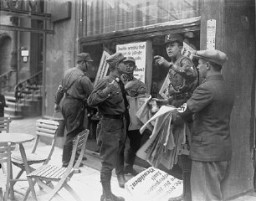 SA üyesi Almanya'da ticarethanelerin yoğun olarak bulunduğu bir caddede nereye Yahudi karşıtı boykot levhalarının asılacağı konusunda diğerlerine talimat veriyor. Sivil giyimli Nazi kolluğu takan Alman genci elinde bir demet Yahudi karşıtı boykot duyurusu tutuyor, diğer SA üyeleri ise duyuruları Yahudi dükkânlarına asıyor. Duyuruların çoğunda “Almanlar kendinizi Yahudi zulüm propagandasına karşı koruyun / Sadece Alman dükkânlarından alışveriş yapın” yazıyor. 1 Nisan 1933 dolayları, Almanya.