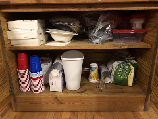 A wooden cabinet full of throw away plastic cups, plastic utensils, paper plates, napkins and other party supplies