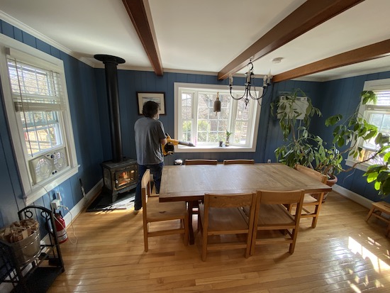 A man in a blue shirt holding a yellow and black Dewalt battery powered leaf blower blowing air to the corner of a dining room.