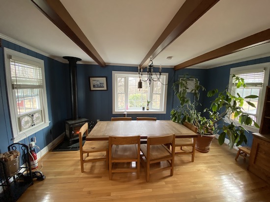 A blue dining room with a This End Up wooden table and chairs in the middle with a wood burning stove and two fans on each end blowing air outside