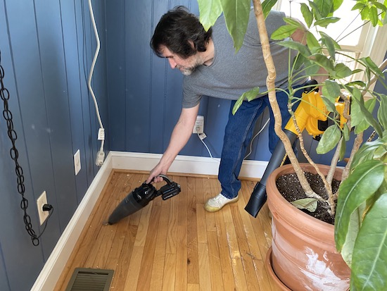 A man with dark hair, a blue shirt and blue jeans sucking up a pile of dirt on a wooden dining room floor