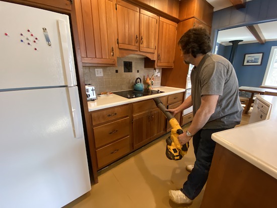 A man in a blue shirt holding a yellow and black Dewalt battery powered leaf blower blowing air in the cracks between a stovetop and the cherry counters below