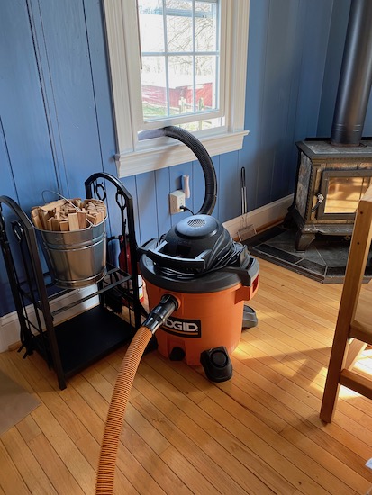 An orange and black shop vac with the exhaust tube out the window next to a wood burning stove