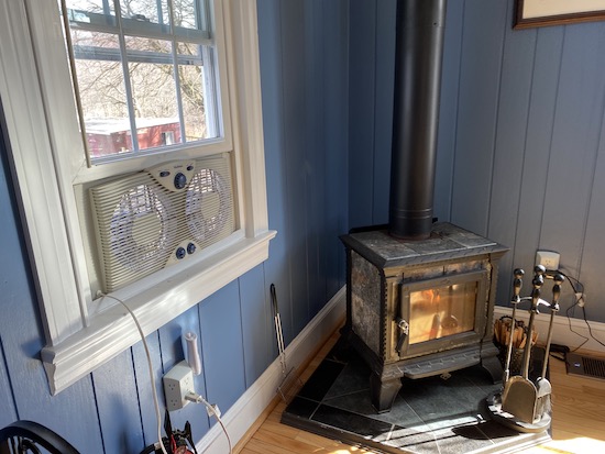 A white exhaust fan blowing air out a window next to a lit wood burning stove