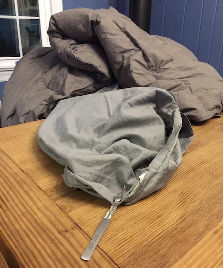 A gray comforter and sheet at a dining room table with a metal seam ripper tool next to it