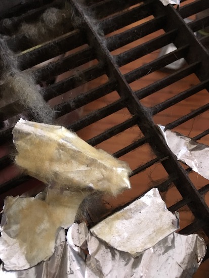 Close up of a black metal air grate with fiberglass stuck to it along with a lot of foil fiberglass backing material